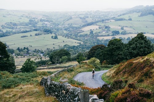 A scenic countryside view with rolling green hills and patchwork fields stretching into the distance. A narrow winding road runs through the foreground, bordered by a stone wall and patches of heather. A cyclist is riding along the road, surrounded by trees and shrubs. The background reveals a valley dotted with farms, hedgerows, and clusters of trees under an overcast sky, creating a tranquil rural atmosphere.