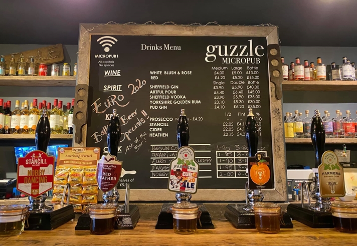 The bar at Guzzle with several beer pumps, a large blackboard with drinks and prices and shelves filled with bottles.