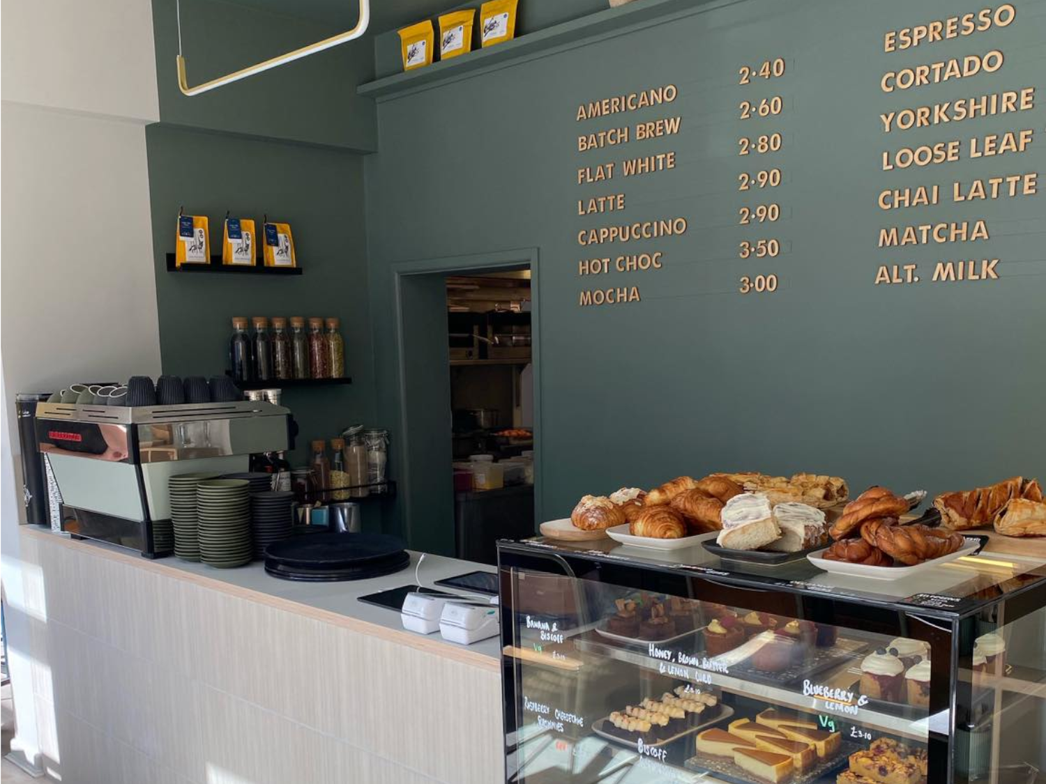 The counter at Kollective Kitchen - Nether Edge with a glass display case full of cakes and pastries. The back wall is a dark sage green with the coffee menu picked out in gold type.