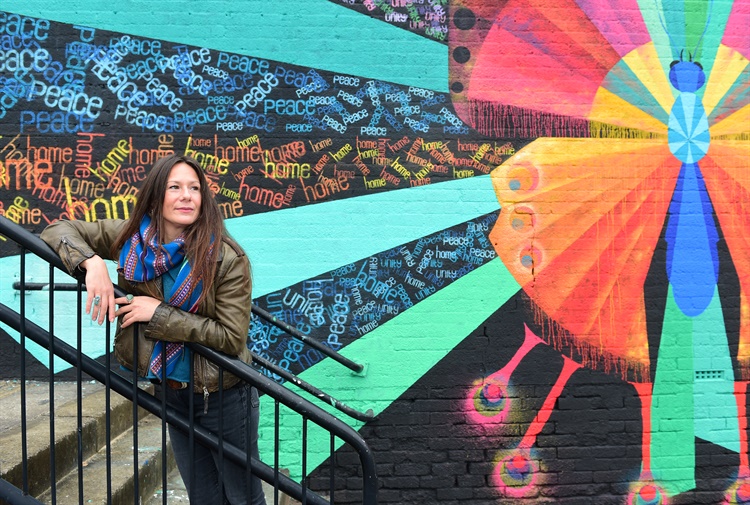 A woman leans on the handrail of some steps, in front of a mural 
