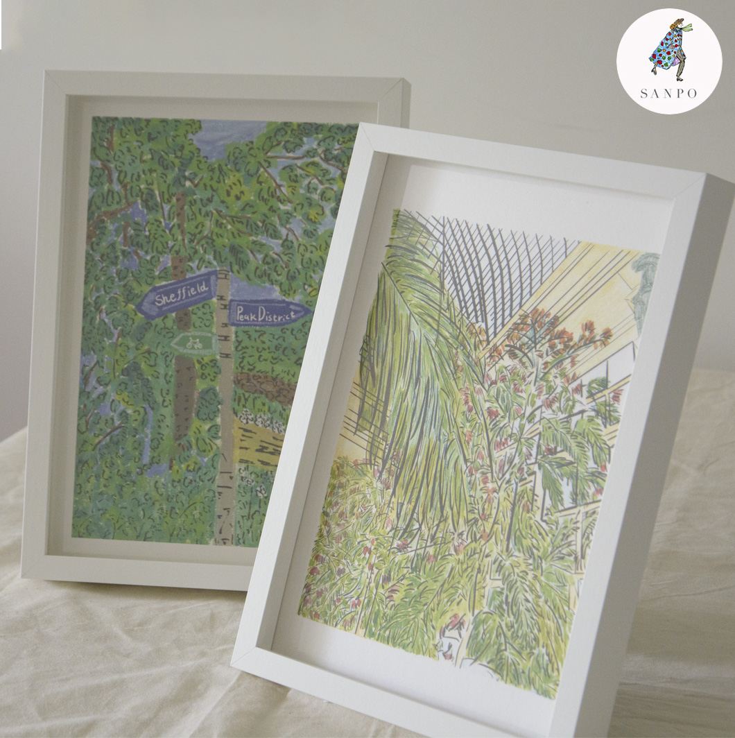 Two framed art prints displayed on a light fabric surface. The left print features a green leafy scene with a signpost pointing to “Sheffield” and “Peak District.” The right print shows a botanical illustration of tall green plants under a glass roof structure, with soft yellow and pink tones in the background.