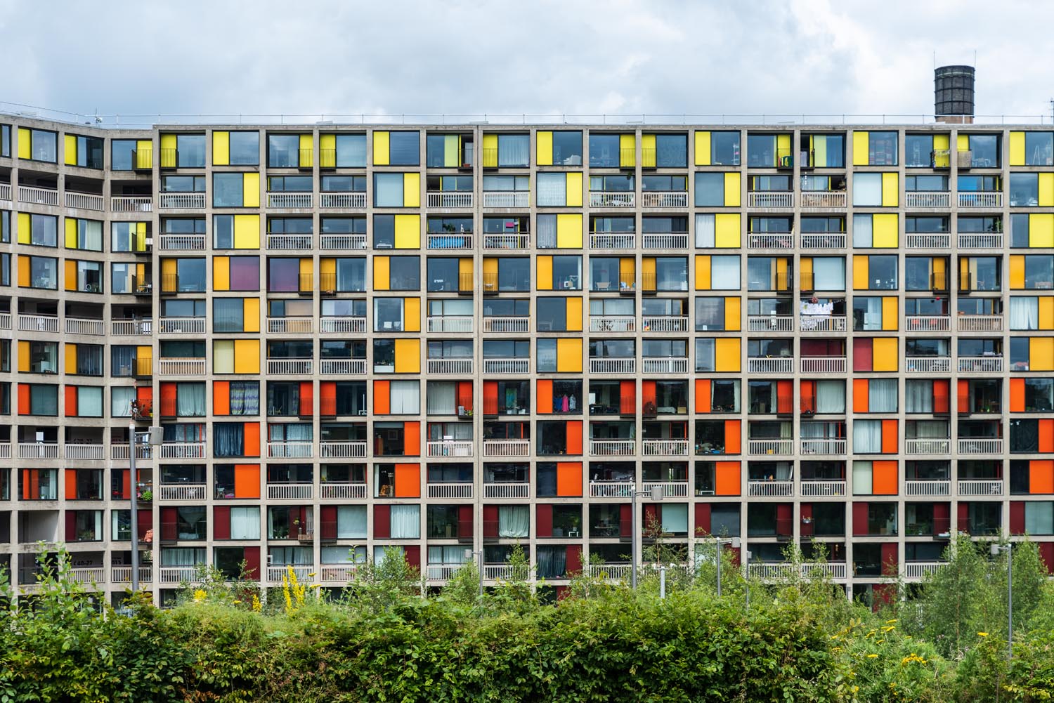 Front view of a large apartment building with a grid-like facade featuring numerous square windows and panels in bright colors, including yellow, orange, and red. The building has multiple balconies and is set against a cloudy sky, with green shrubs and trees in the foreground.