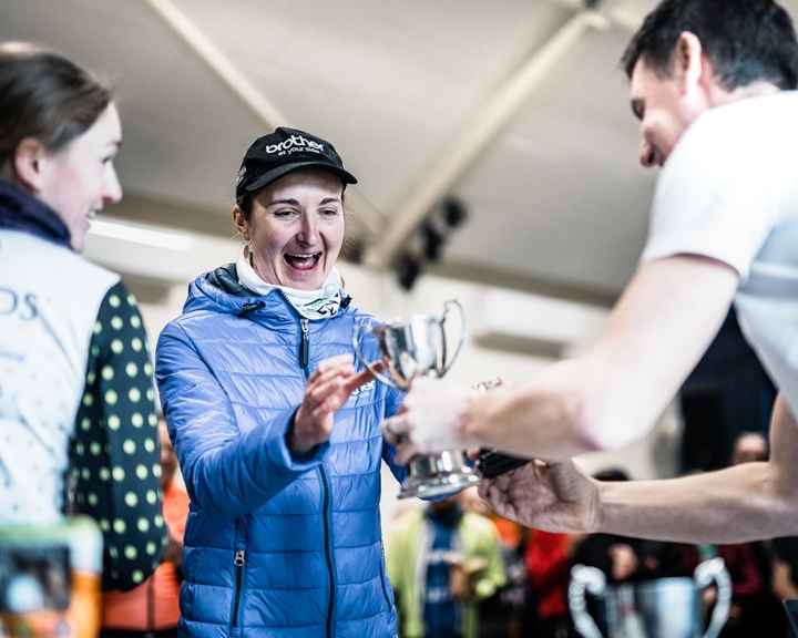 Woman excited as she receives a small silver trophy being handed to her by a man. Another female is also in conversation to the left.