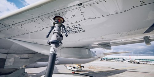 Close-up view of an aircraft wing during refueling at an airport. A fuel hose is connected to the underside of the wing through a metal coupling. The wing surface shows visible rivets, panels, and printed technical markings. In the background, airport ground vehicles and terminal buildings are visible under a partly cloudy sky.