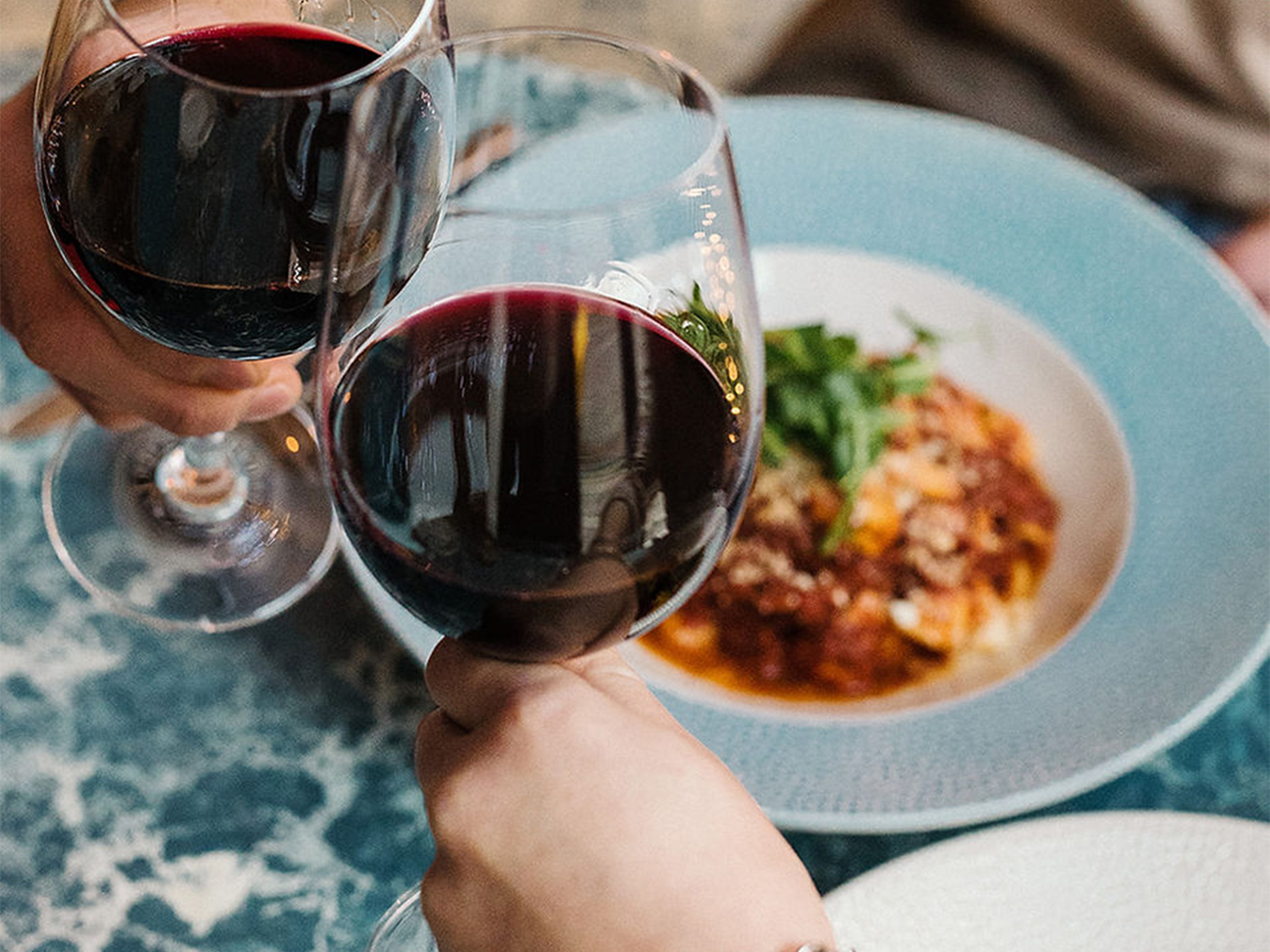 Two wine glasses clinking above plates of food.