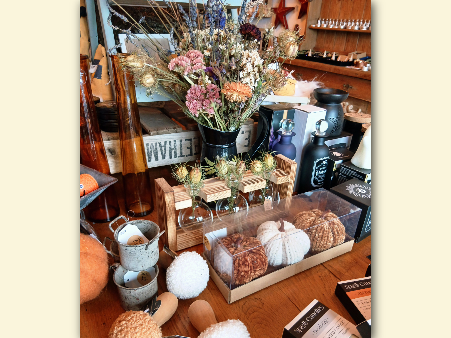 A tabletop display of rustic home décor items, including a black vase filled with dried flowers in shades of pink, purple, and beige, a wooden holder with three small glass vases containing dried thistles, and a clear box with textured decorative pumpkins in white, brown, and tan. Surrounding items include small metal cups, fluffy pom-poms, amber glass bottles, and boxed candles, all arranged in a cozy, autumn-themed setting.