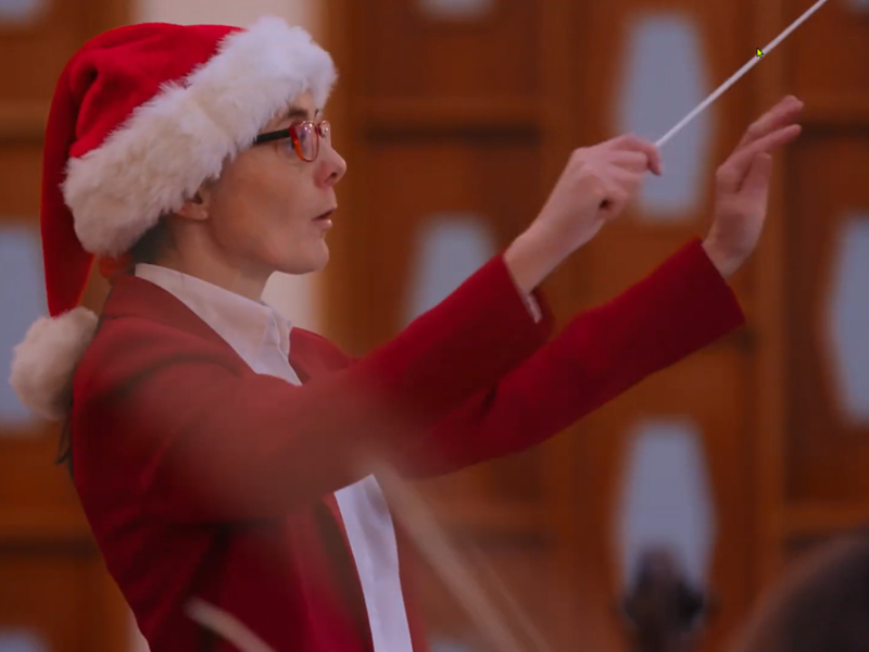 A woman in a red outfit and a Santa hat conducts an orchestra.