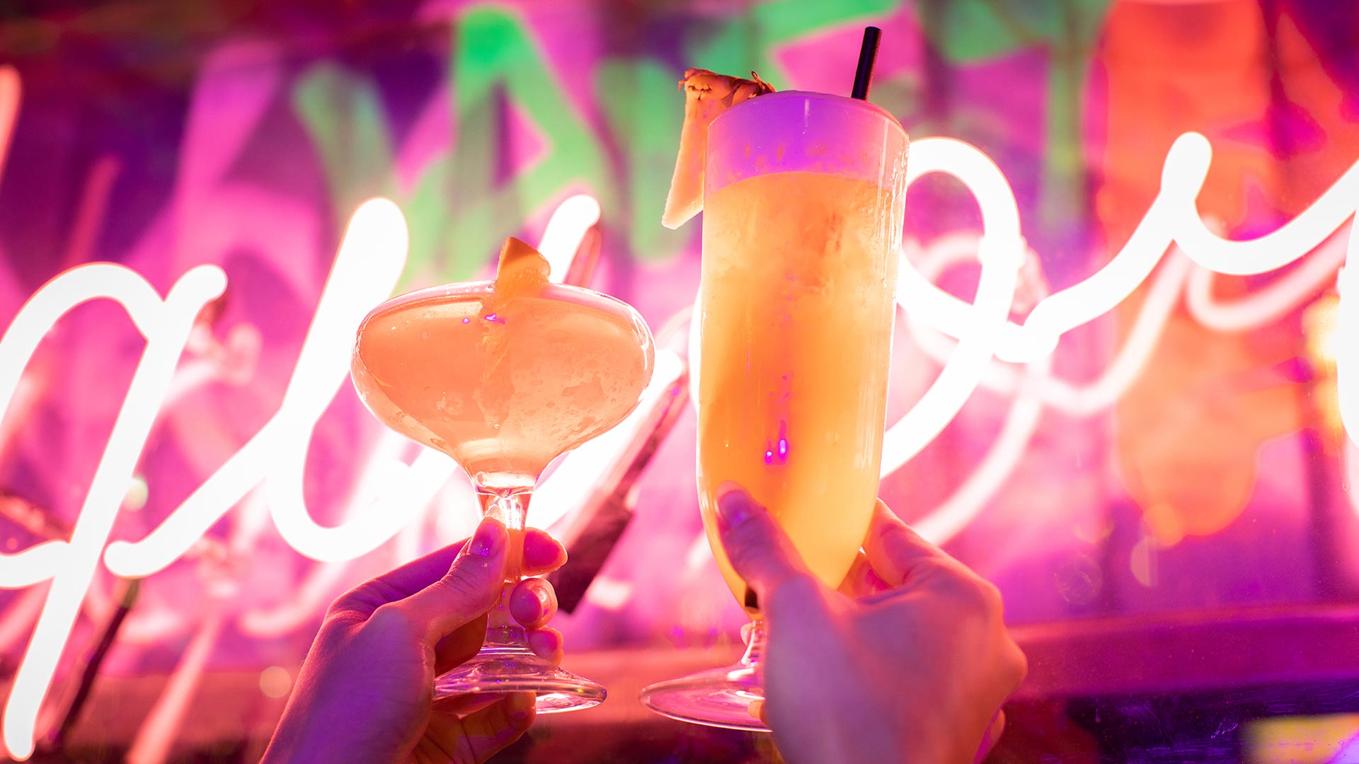 Two cocktails being held up in the in front of a neon sign.