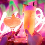 Two cocktails being held up in the in front of a neon sign.