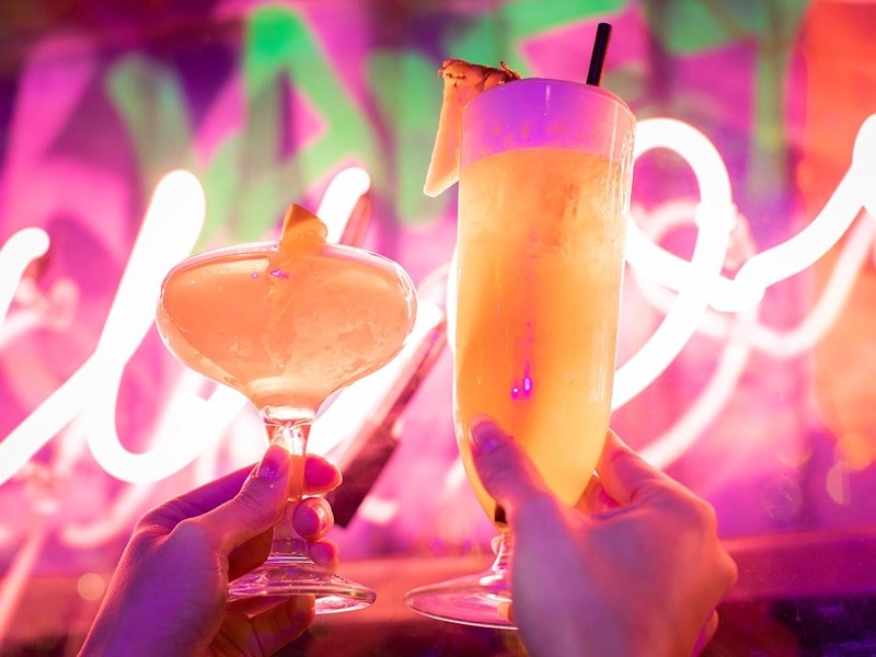 Two cocktails being held up in the in front of a neon sign.