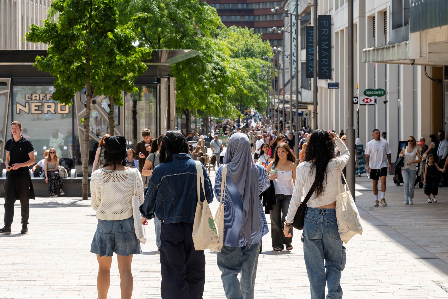Four people walking down a busy pedestrian shopping street lined with stores and outdoor seating. Visible shop signs include “Caffè Nero” on the left and “Primark” on the right. The street is shaded by green trees, and the background shows a large crowd of people, modern buildings, and bright daylight.