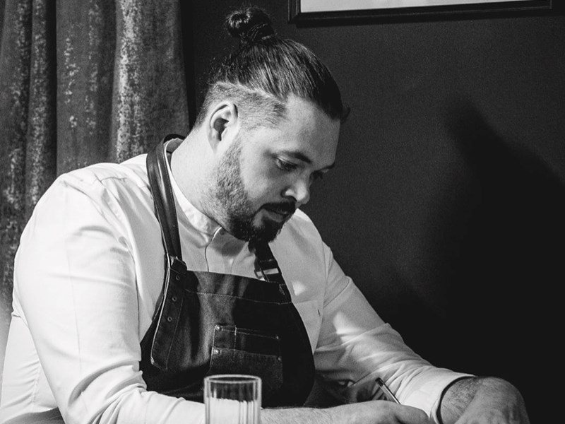 A black and white picture of a man in chef's clothing, writing at a tabel.