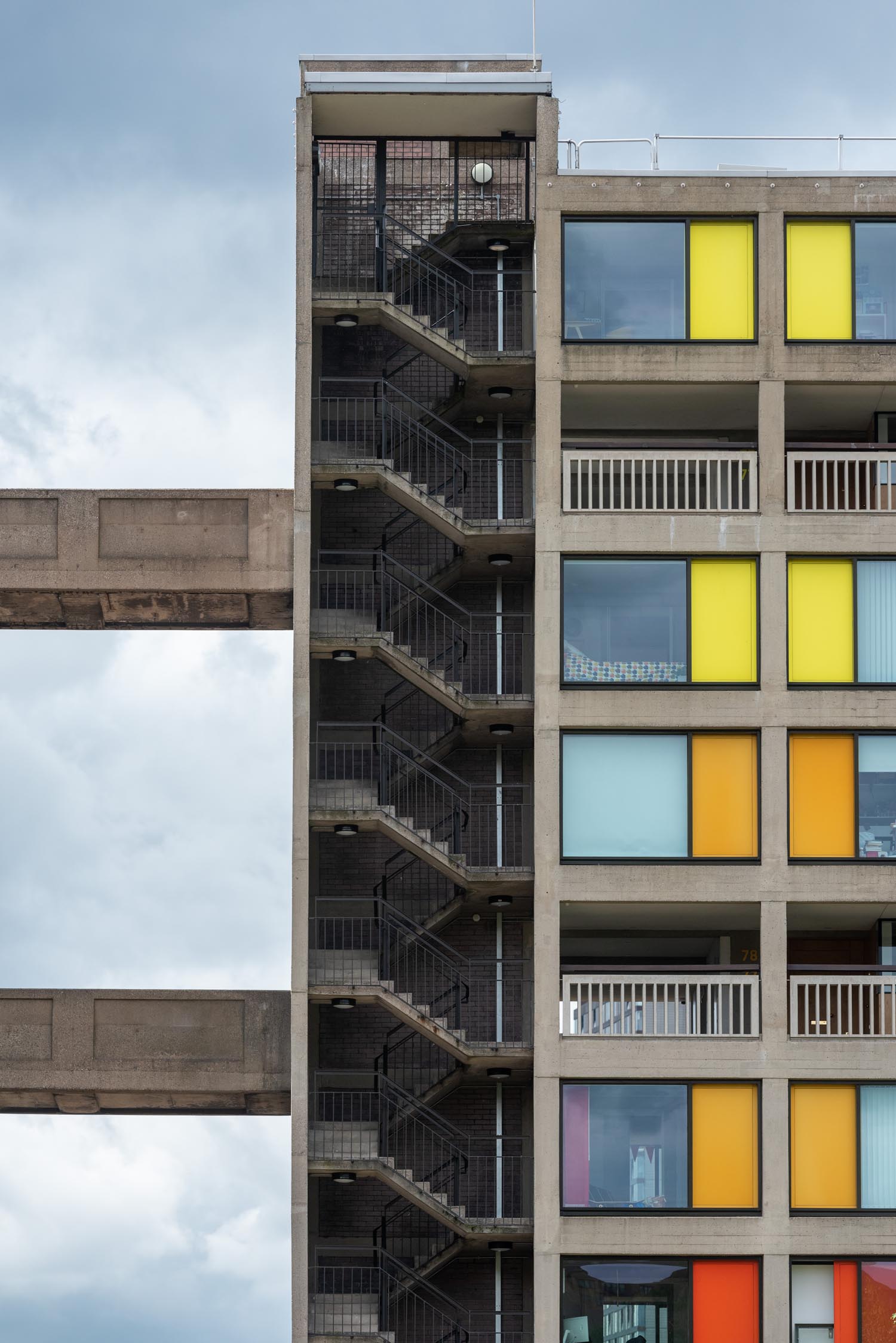 Close-up view of a modern concrete apartment building with an external zigzag staircase on the left and colorful window panels on the right in shades of yellow, orange, and blue. The structure includes connecting walkways and balconies, set against a cloudy sky.