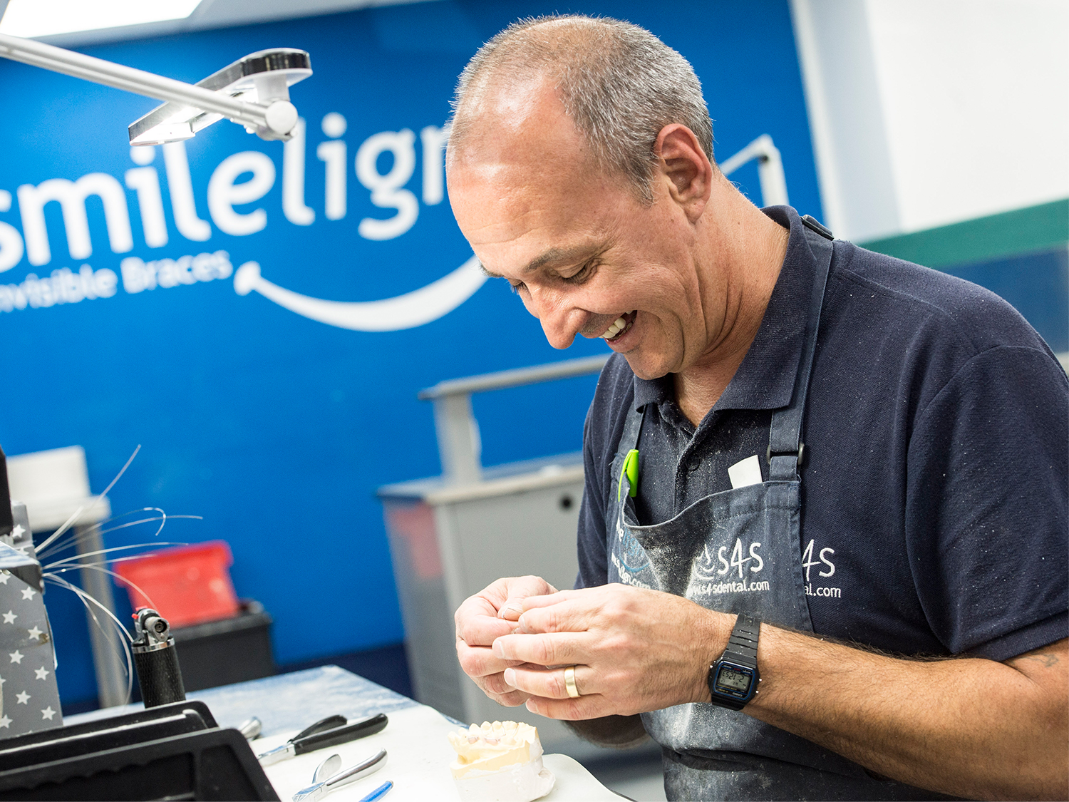A smiling male technician, in work-wear and an apron, sits working on a set of hi-tech braces for the dental sector.