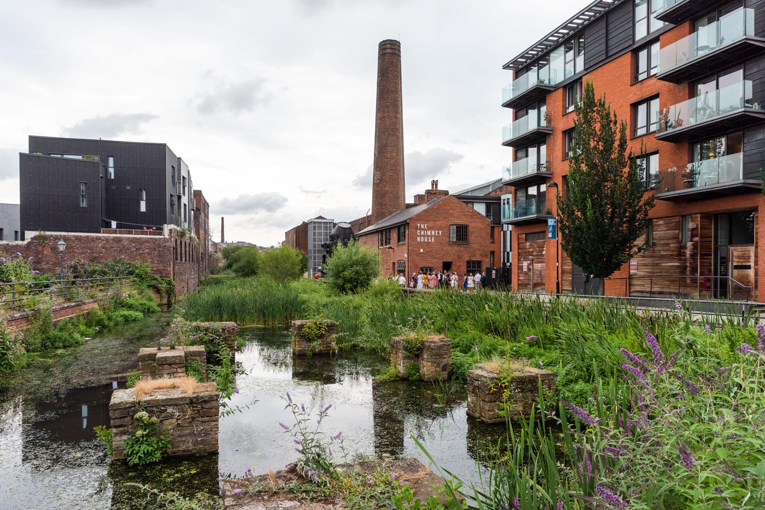 A scenic view of a canal surrounded by greenery and historic architecture. The water reflects the sky and nearby plants, with remnants of old stone structures partially submerged. On the right, modern red-brick and glass buildings line the canal, while a tall industrial chimney rises prominently in the background. A brick building labelled “The Chimney House” is visible near the centre, and purple wildflowers grow along the water’s edge.