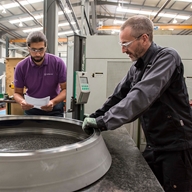 Two men working in an industrial setting. Both are wearing safety glasses and one of them is reading a sheet of paper.