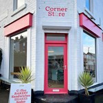 A brick building that has been painted white, with red window frames and door. There is a hand painted sign, in red, above the door that reads 'Corner Store'. The letter 'o' in the word Store is a smiley face.