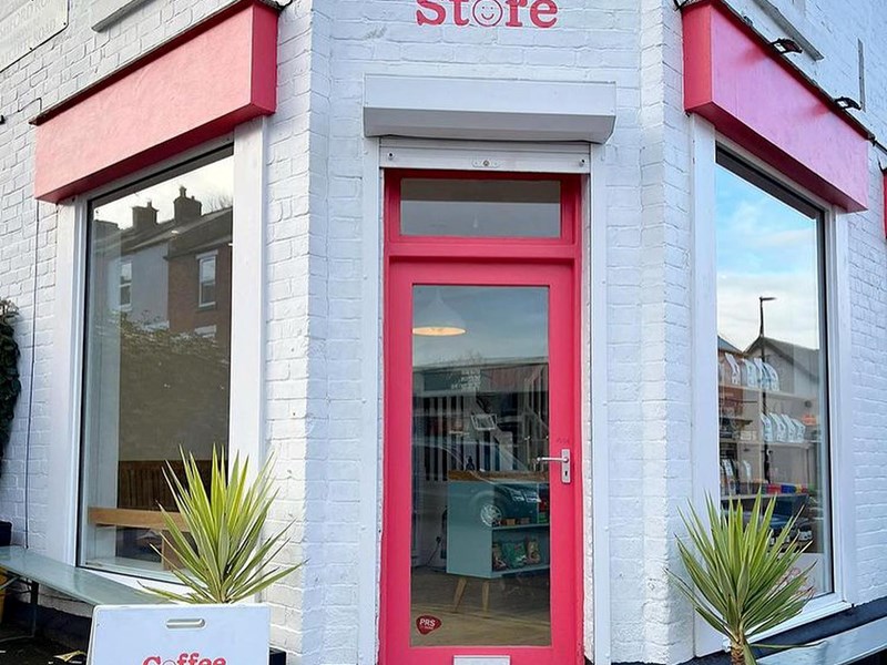 A brick building that has been painted white, with red window frames and door. There is a hand painted sign, in red, above the door that reads 'Corner Store'. The letter 'o' in the word Store is a smiley face.