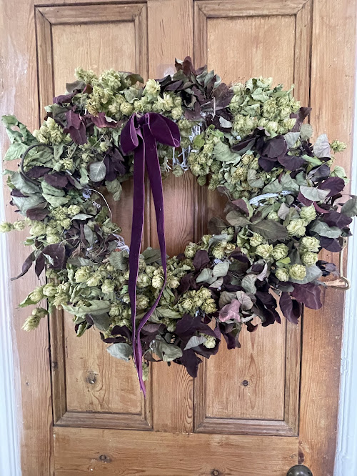 A festive wreath made from dried green hops and deep burgundy leaves, creating a textured and vibrant display. A purple velvet ribbon hangs from the center, and the wreath is mounted on a wooden door.