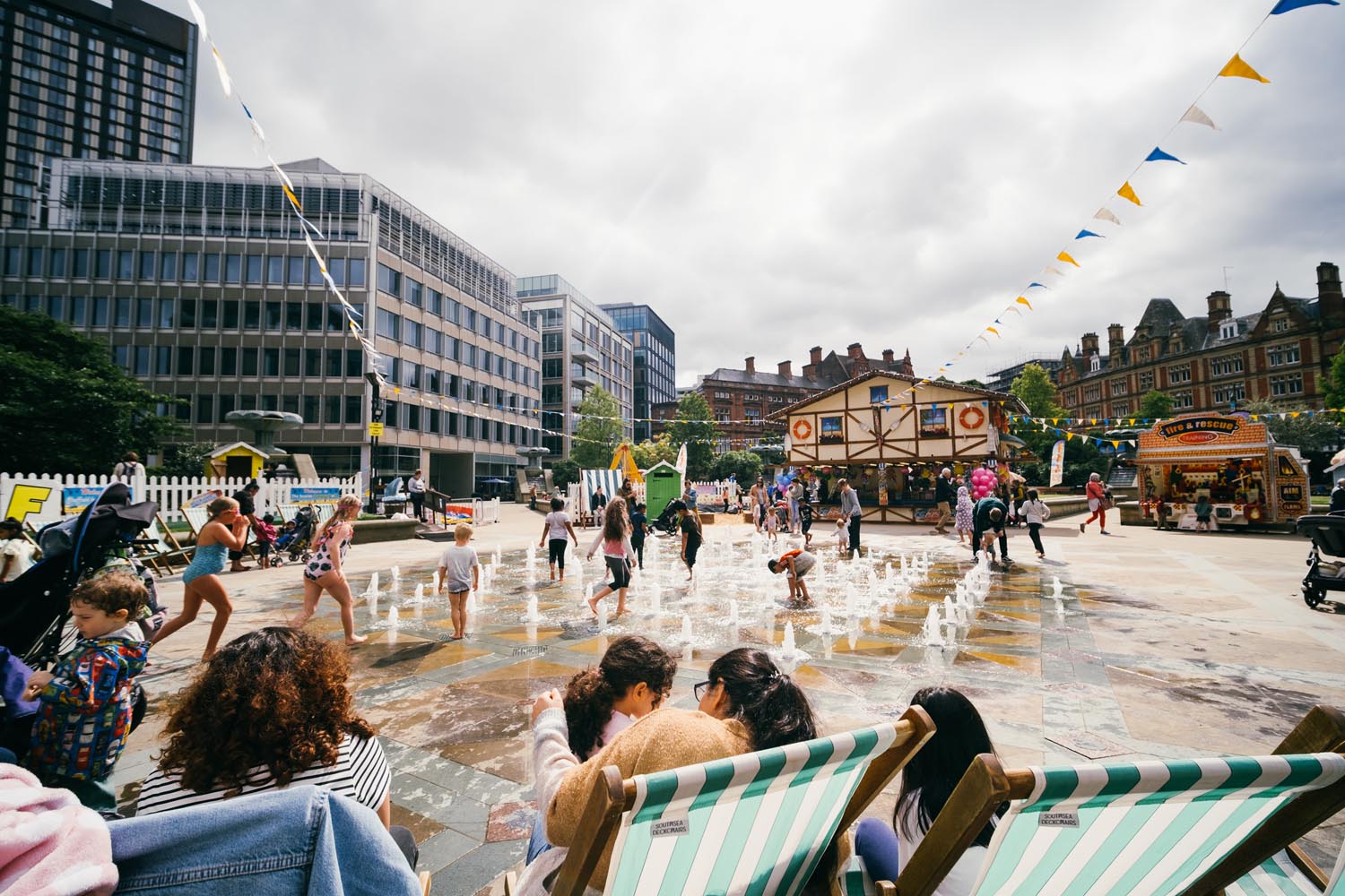 Children play in a city centre fountain, with adults sat around the perimeter in deck chairs.