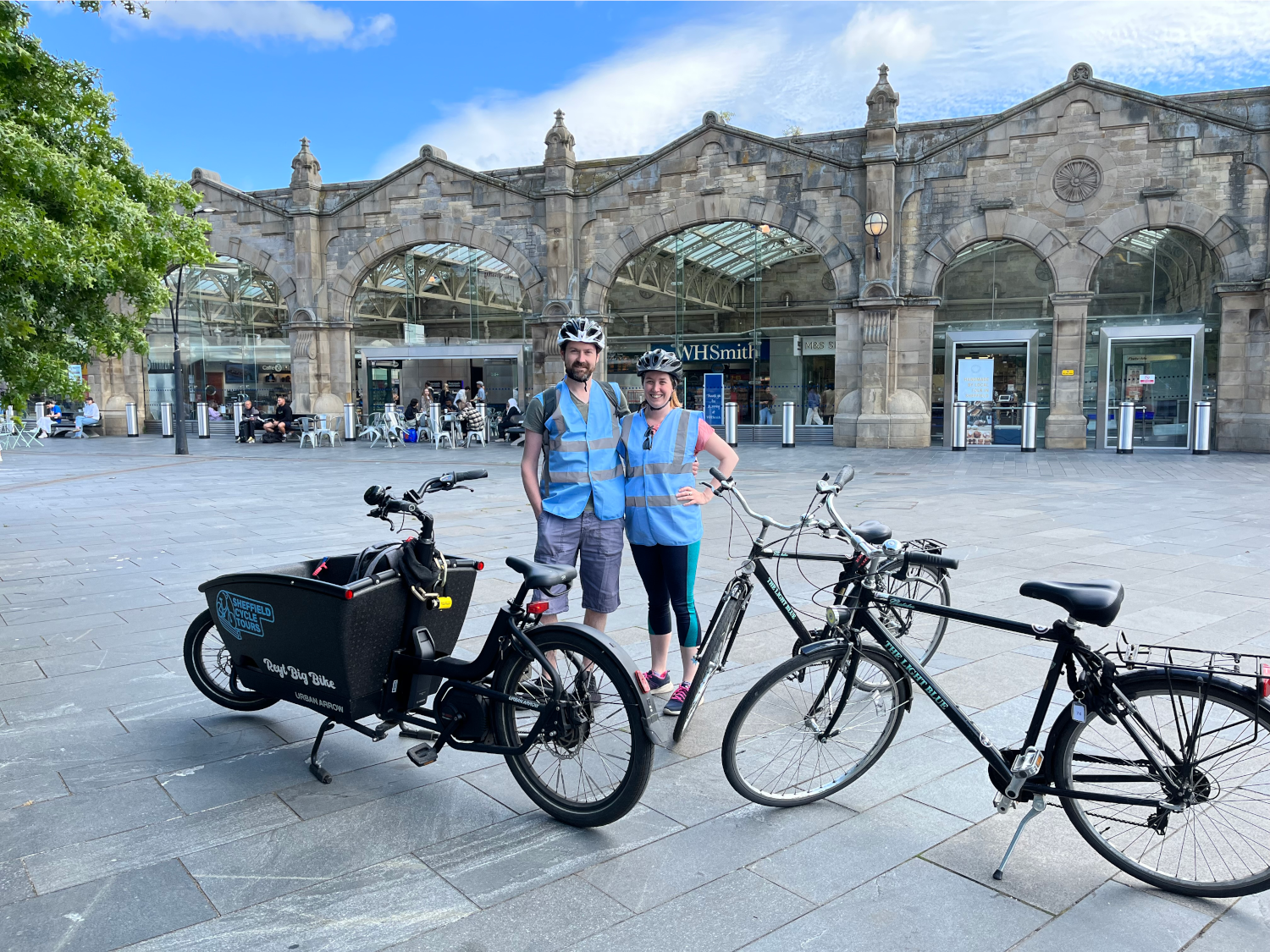 Sheffield Cycle Tours - Two people wearing blue safety vests and helmets standing with bicycles in front of a historic stone building with arched glass entrances. The building has signage for WHSmith, and the foreground shows a cargo bike and two standard bicycles on a paved plaza under a partly cloudy sky.