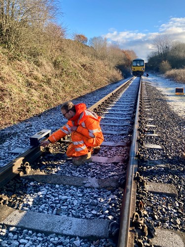 Railway maintenance worker in bright orange high-visibility gear kneeling on a frosty track, adjusting equipment attached to the rail. A train is visible in the distance on the same track, surrounded by trees and bushes under a clear blue sky.