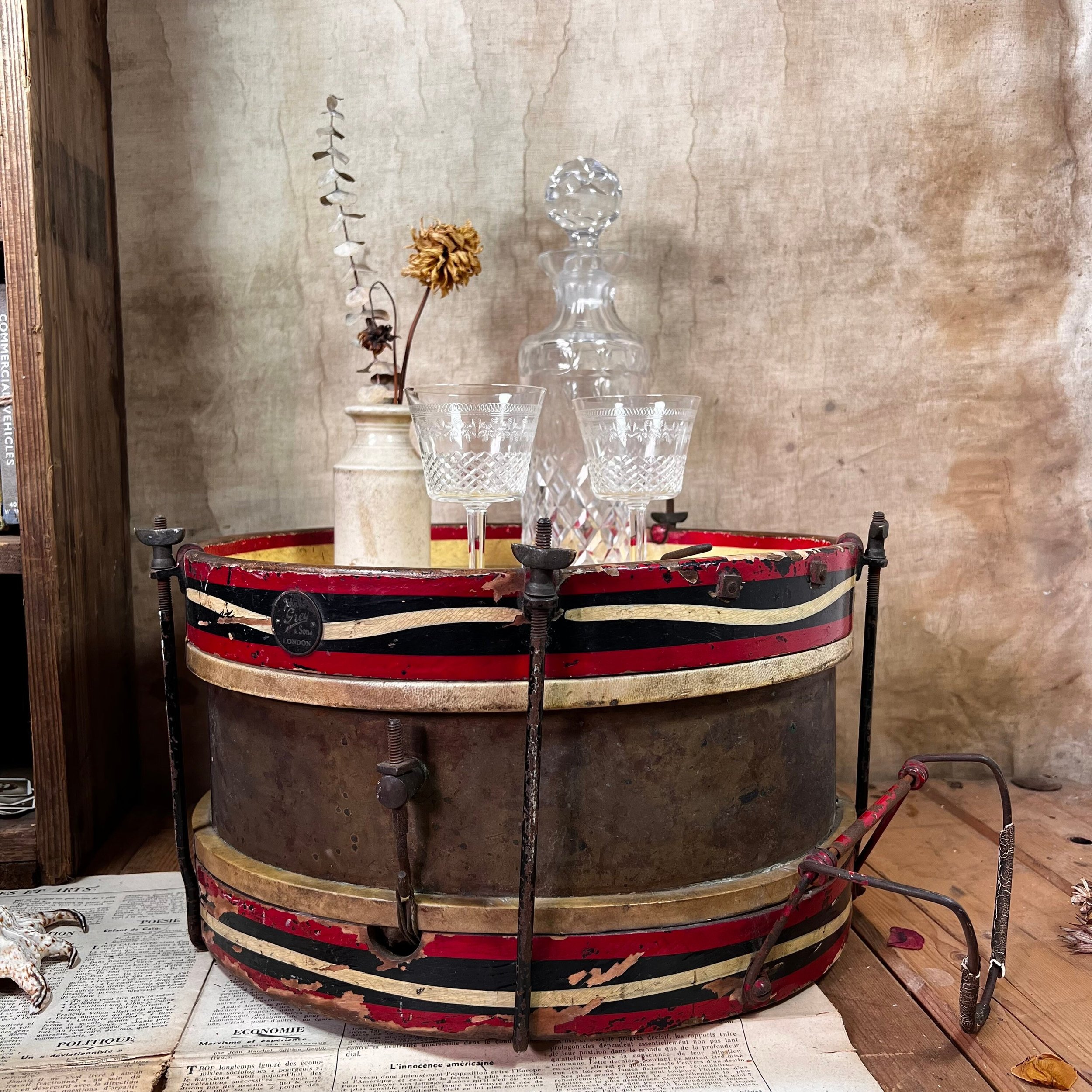 A bass drum lying on its side, on the floor, with a vase of flowers, a decanter and two glasses on top.