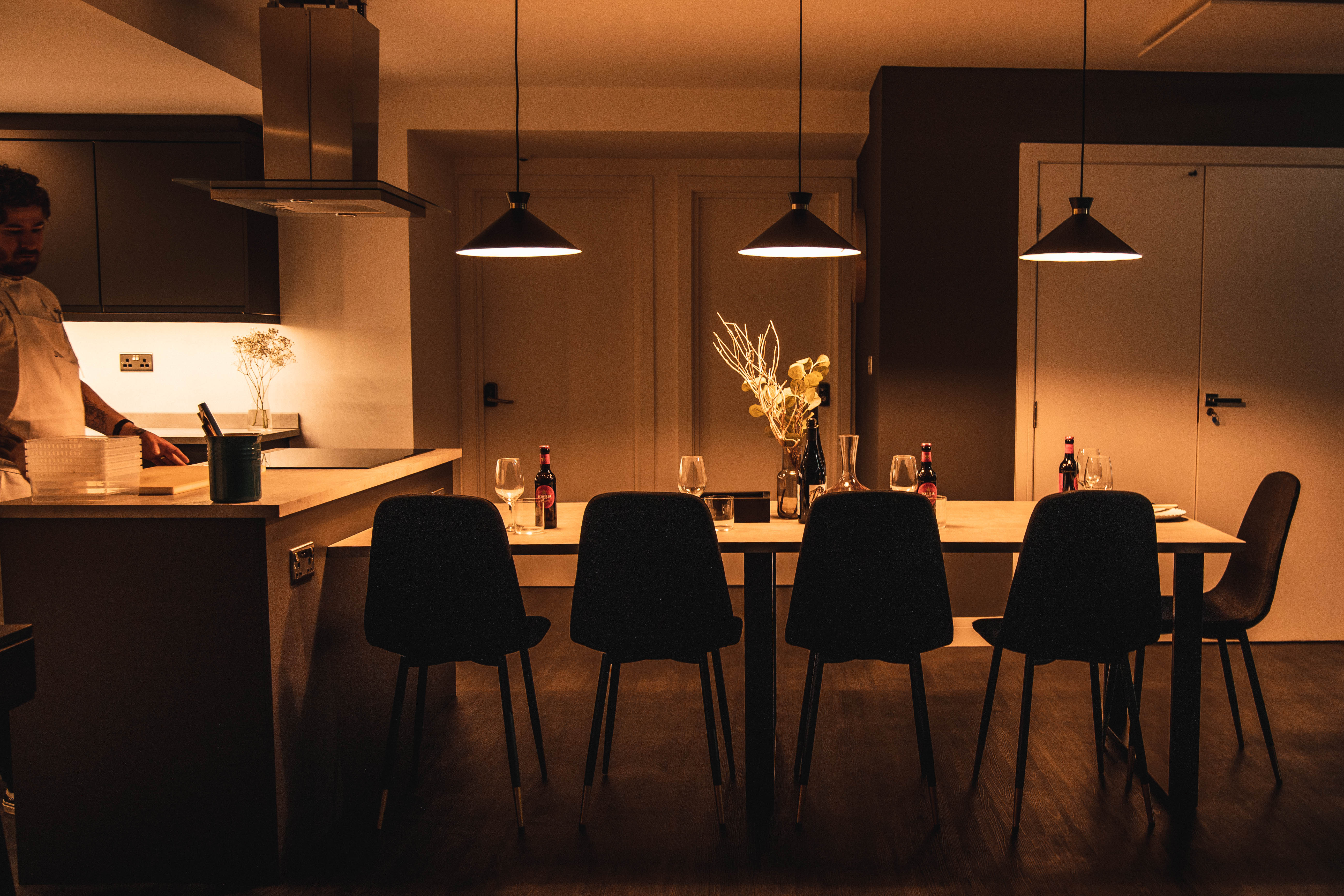 A chef stands at a counter, next to a table laid out for five people. The scene is lit by subdued lighting.