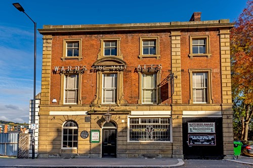 Historic three-story brick building with stone detailing and large windows, featuring signage that reads ‘WARD’S FINE MALT ALES.’ A black banner on the right advertises a ‘RATTLER AWARD.’ The building is set on a street corner with a lamppost and autumn trees in the background under a clear blue sky.