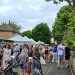 A crowd of people walking along the Nether Edge Farmers' Market. There are stalls situated in front of houses to the left, while the people walk alongside it to the right.