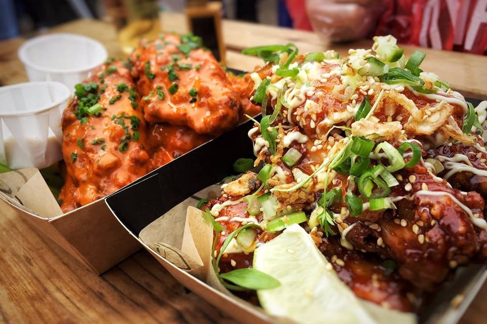 Close-up of two trays of saucy chicken wings served on a wooden table. The tray in the foreground features wings coated in a sticky glaze, topped with sesame seeds, chopped green onions, and a drizzle of creamy sauce, with a lime wedge on the side. The tray in the background contains wings covered in bright orange sauce, garnished with finely chopped herbs. Two small white dipping cups are placed alongside the wings.