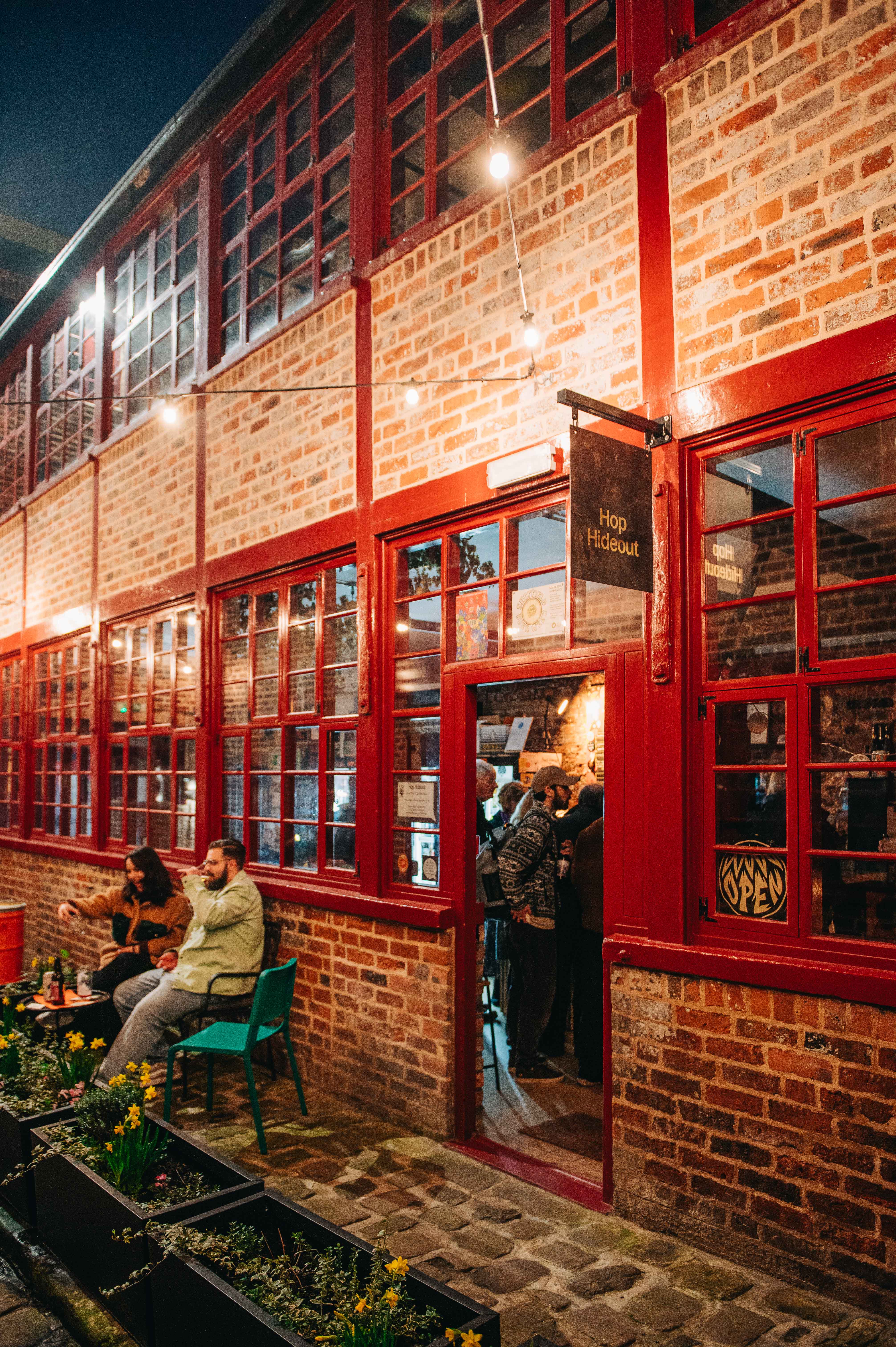 The red brick exterior of Hop Hideout at Leah's Yard, with red painted windows and doors and a black hanging sign that says 'Hop Hideout'. A man and a woman are sat on a table outside drinking, the door is open and customers are waiting at the bar.