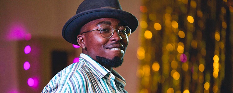 A man of African heritage, wearing a pork pie hat and a striped shirt, smiles broadly.