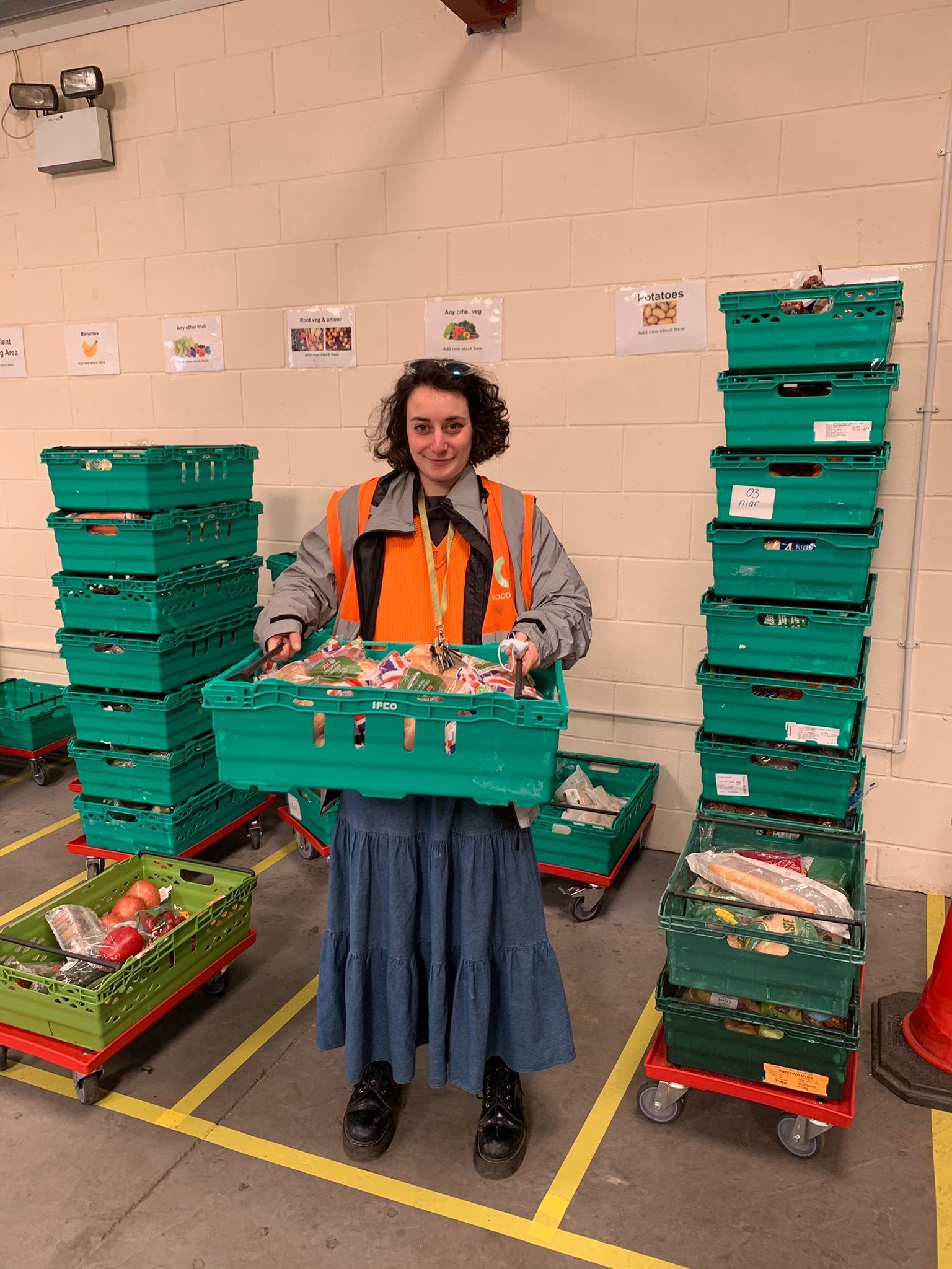 Person wearing a gray jacket and orange high-visibility vest, holding a green plastic crate filled with assorted packaged food items. The individual is standing in a warehouse-like setting with beige walls, yellow floor markings, and multiple stacks of green crates on wheeled trolleys arranged around them.