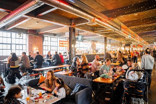 A lively indoor food hall with long wooden tables and benches filled with people eating and drinking. The space has exposed brick walls, large industrial-style windows, and string lights hanging from the ceiling, creating a warm and festive atmosphere. A stroller is visible in the foreground, and the hall extends into the background with more diners.