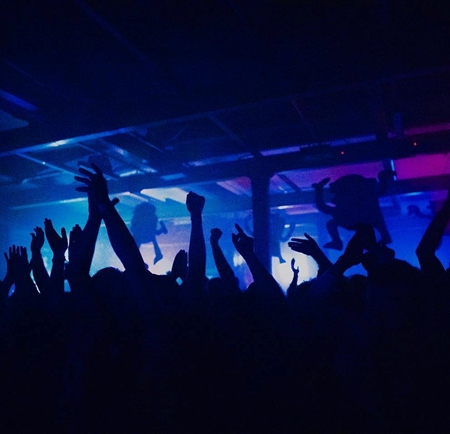 A crowd dances, arms held high, at a night club. The lighting is all blues and purples.