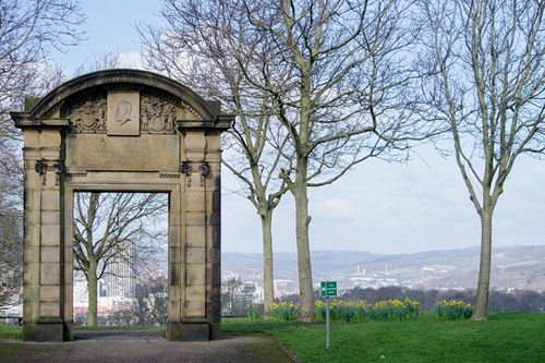 A historic stone archway with decorative carvings stands in a park surrounded by bare trees. The arch frames a distant view of a cityscape with hills in the background. Yellow daffodils bloom in the grassy area near the arch, and a small green sign is visible on the lawn. The sky is bright with scattered clouds, creating a clear and open atmosphere.