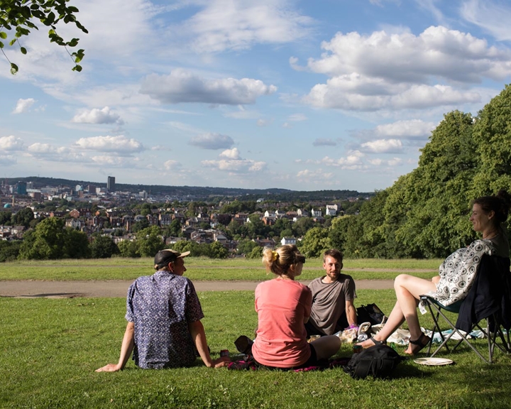 A group of four people, sat in Meersbrook Park, enjoying a sunny day. One is sat in a folding chair and the other three are sat on the grass.