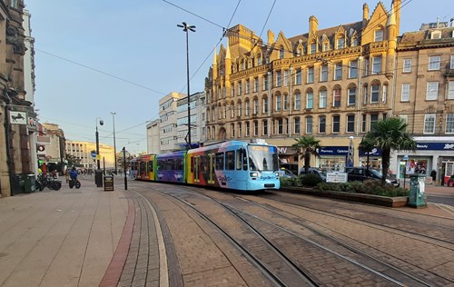 Street scene featuring a colorful tram traveling along tracks in a city center. The tram is painted in bright blue and multicolored designs. Surrounding architecture includes ornate historic buildings with large windows and decorative stonework. Palm trees and planters line the street, and pedestrians are visible in the background under a clear blue sky.