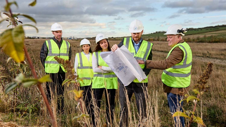 Five people in high viz jackest and hard hats stand in a field studing some plans.