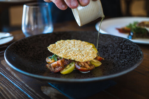 A close-up of a dark textured plate containing a colorful dish of sliced heirloom tomatoes topped with a large, crisp parmesan tuile. A hand is pouring a light green sauce or dressing from a small white jug onto the dish. In the background, there is a glass of water and another plate with food, slightly out of focus, on a wooden table.