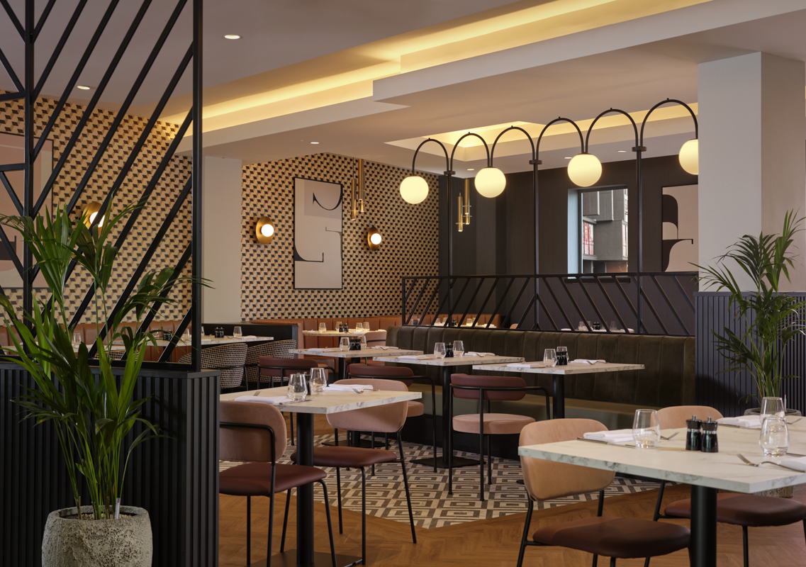 Modern restaurant interior with marble-top tables, cushioned chairs in muted tones, and geometric patterned flooring. The space features warm ambient lighting, decorative wall panels with abstract art, and a row of globe pendant lights above a booth seating area. Green potted plants add a touch of nature.