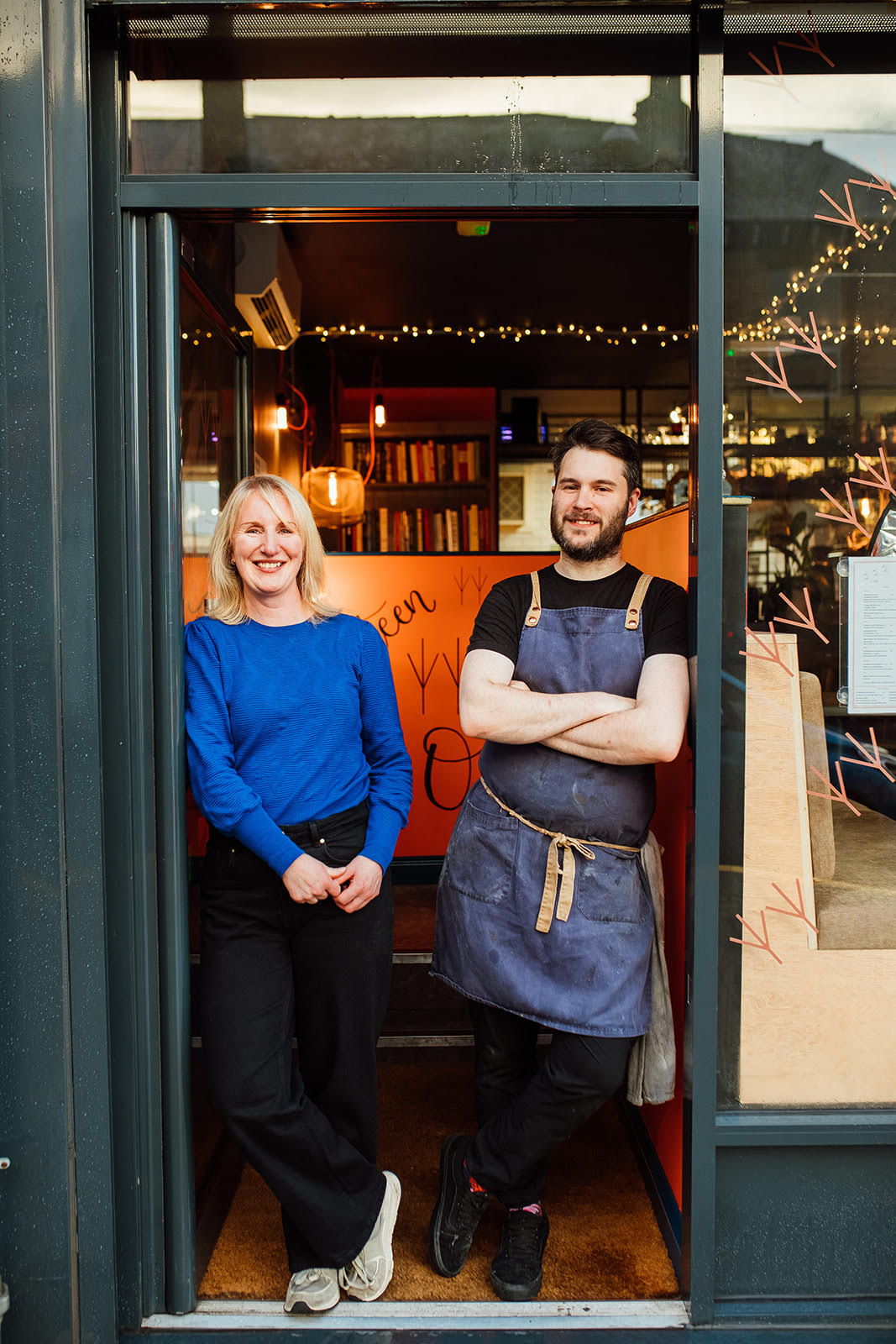 Two people standing in the doorway of a cozy restaurant or café. One is wearing a bright blue long-sleeve top and dark pants, while the other is dressed in a black shirt with a dark apron tied at the waist. The interior behind them features warm lighting, shelves with books, and string lights. A handwritten orange sign is partially visible inside, and the exterior has dark framing around the entrance.