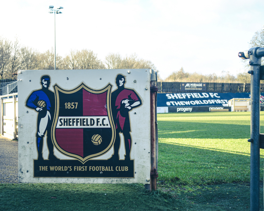 Signboard featuring the Sheffield F.C. crest with text ‘1857 Sheffield F.C.’ and ‘The World’s First Football Club,’ flanked by illustrations of two footballers. In the background, a football pitch and advertising boards display ‘Sheffield FC #TheWorldsFirst.’ Trees and sky are visible beyond the stands.