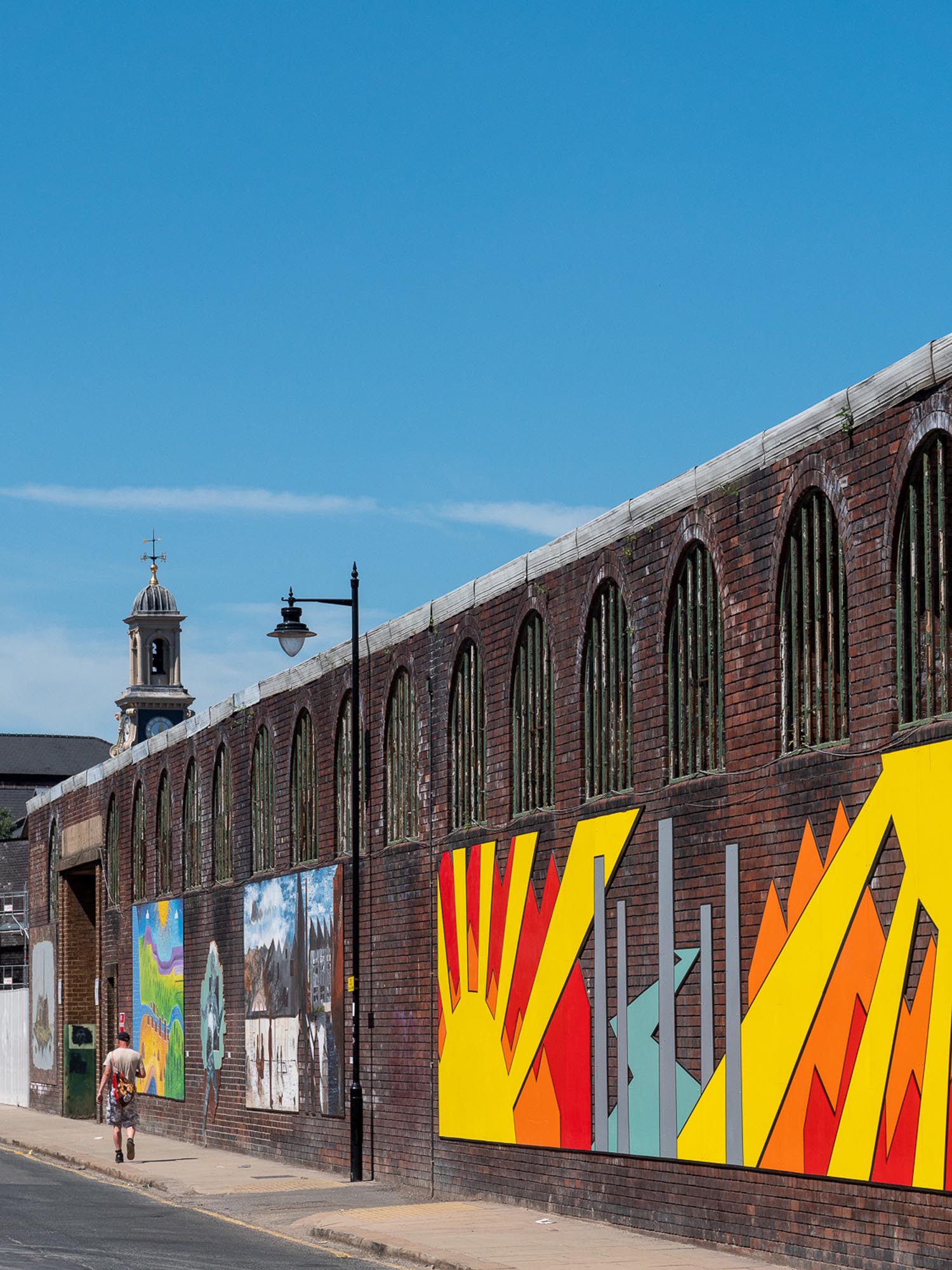 A long brick wall with arched windows features a series of colorful murals, including abstract geometric designs in red, yellow, orange, and gray. Additional artworks with varied styles are visible along the wall. A person walks on the sidewalk beside the wall, and a historic building with a clock tower rises in the background under a clear blue sky.