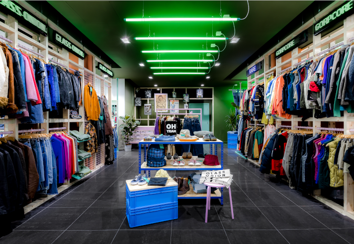 Inside the Glass Onion shop. All the walls are full of clothes on rails. In the centre of the shop are tables with accessories on display.