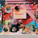 A woman is sat smiling behind a stall, selling knitted and crocheted goods.
