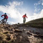 Two people mountain biking down a slope out in the countryside.