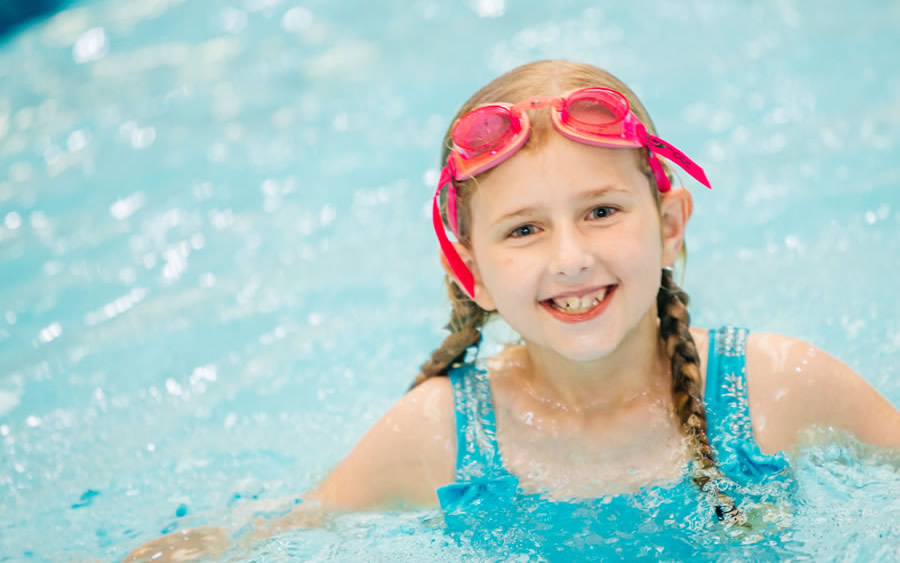 Child wearing a turquoise swimsuit and pink swimming goggles, partially submerged in a clear blue swimming pool with rippling water.