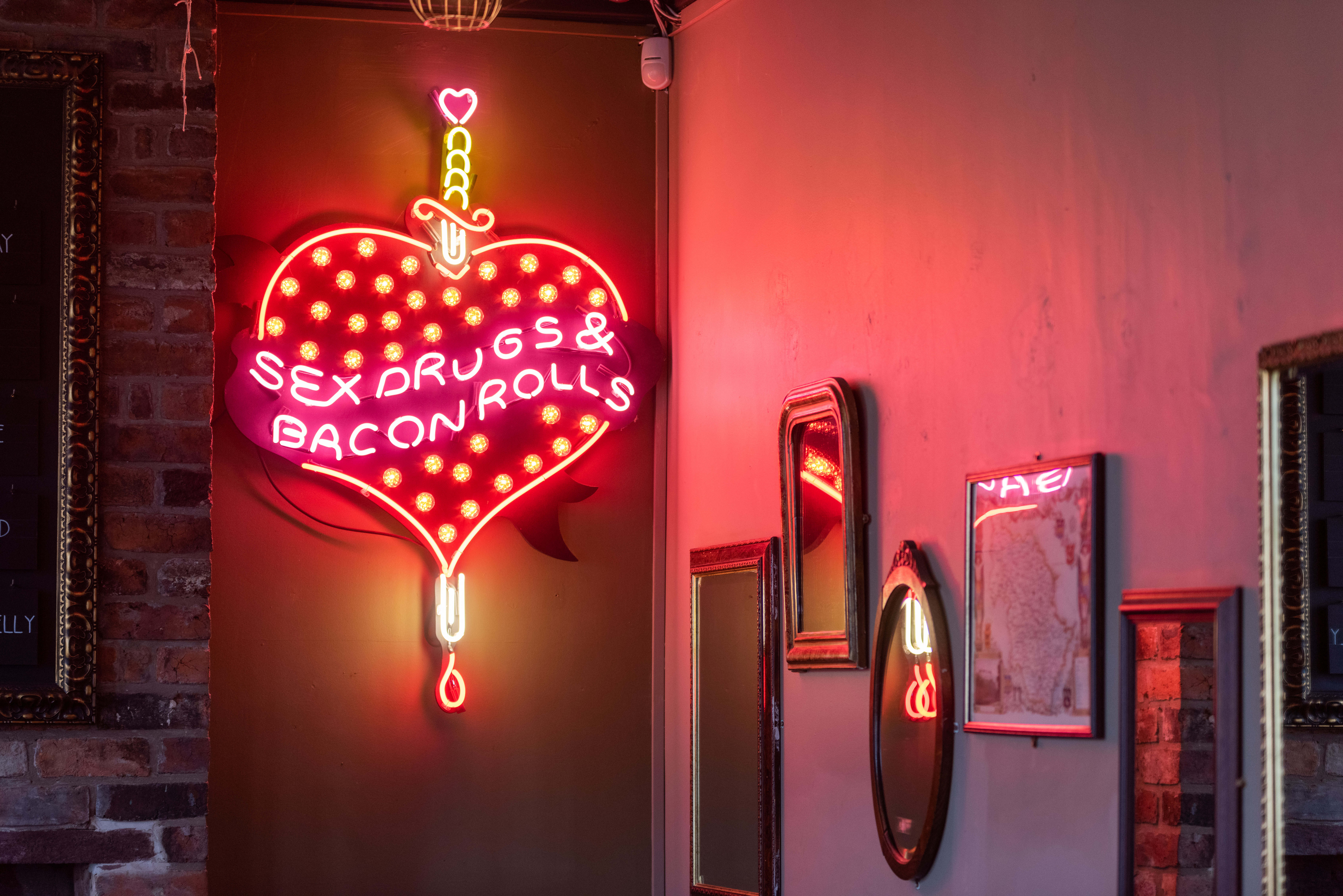 A cheeky neon sign that shows a heart pierced by a dagger with the words 'Sex, drugs & bacon rolls' on the wall at The Riverside Kelham.
